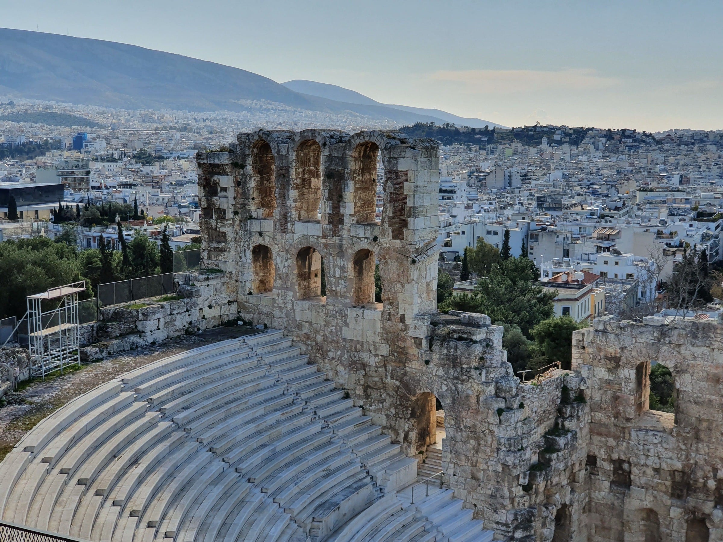 The Odeon of Herodes Atticus below the Acropolis in Athens, the city stretching behind it under a soft sky