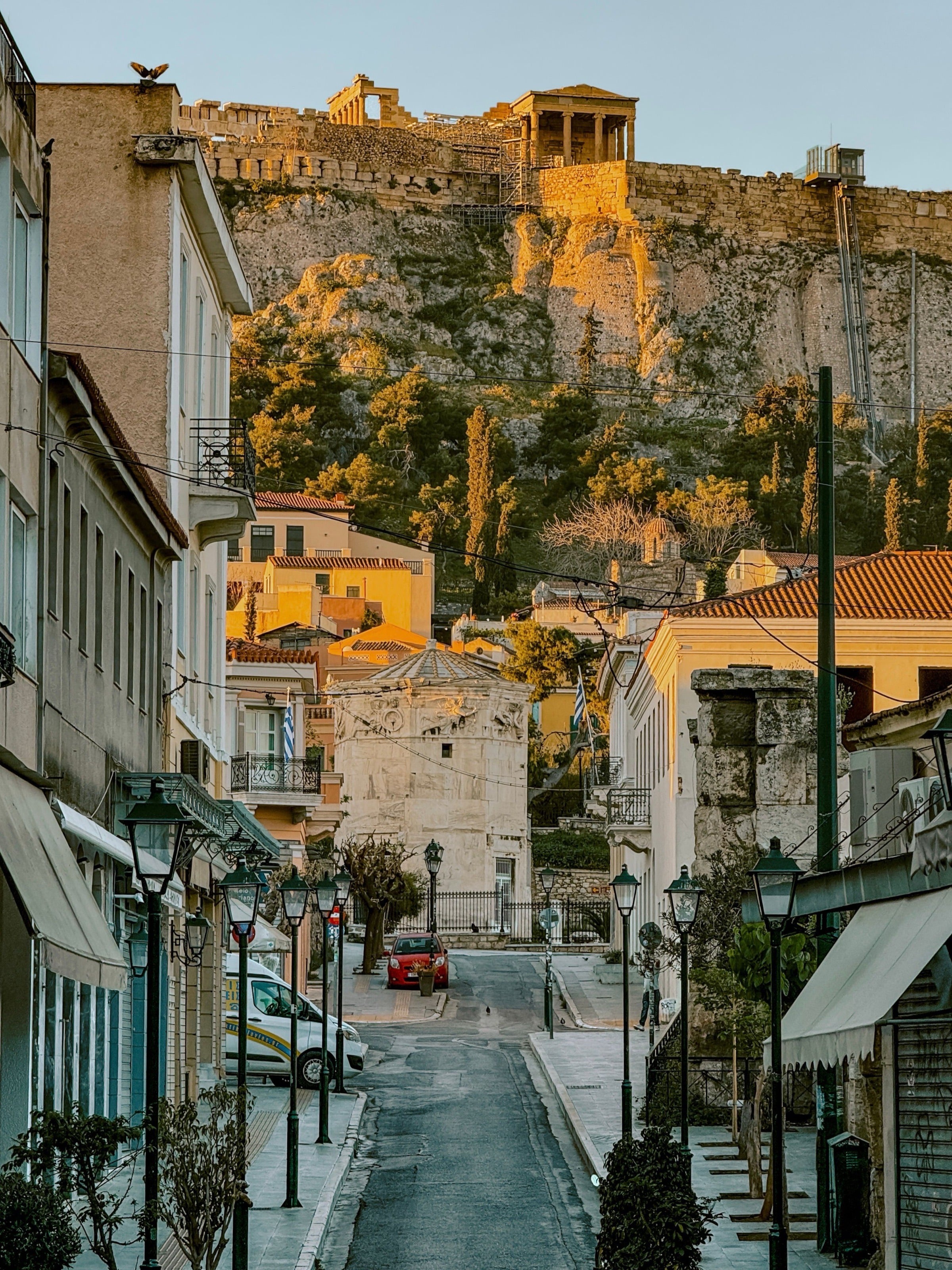 Dusk in Plaka — golden light on the Acropolis seen from a quiet street below, the Roman Tower of the Winds in the middle distance.