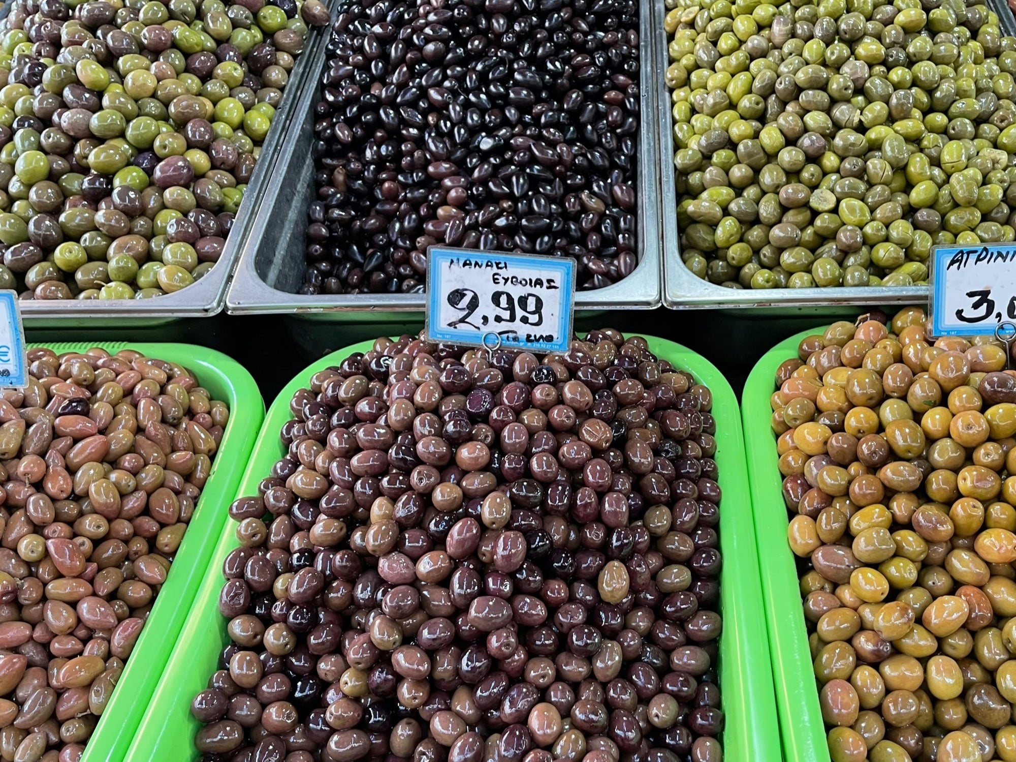 Trays of green, black and violet olives on a market counter in central Athens, each with a handwritten Greek price card.