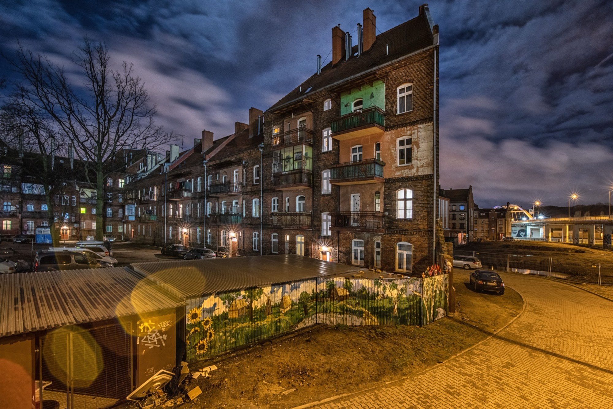 A quiet residential street in Gdańsk at twilight — weathered brick tenements with lit windows, a painted hoarding and a near-empty road.