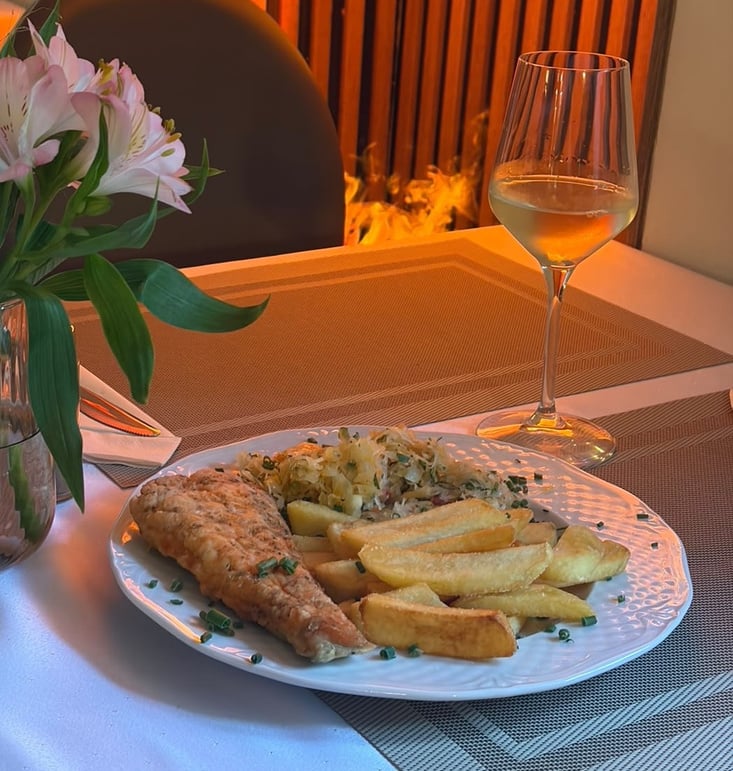 Pan-fried salmon with steakhouse-style chips and a glass of wine on Restauracja Celestin's table, the dining room's fire glowing behind