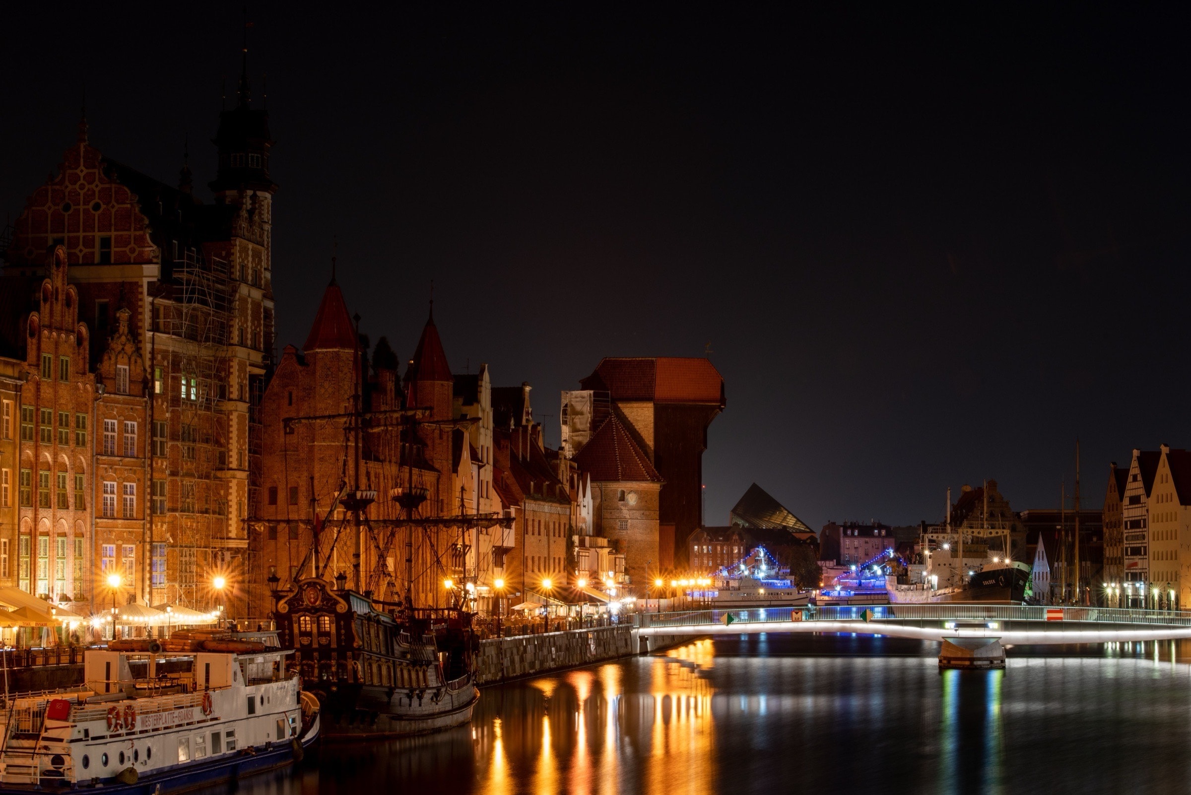 The Długie Pobrzeże waterfront and the Żuraw crane lit warmly after dark, lamplight reflected in the Motława — one of the streets the fair's stalls run along.