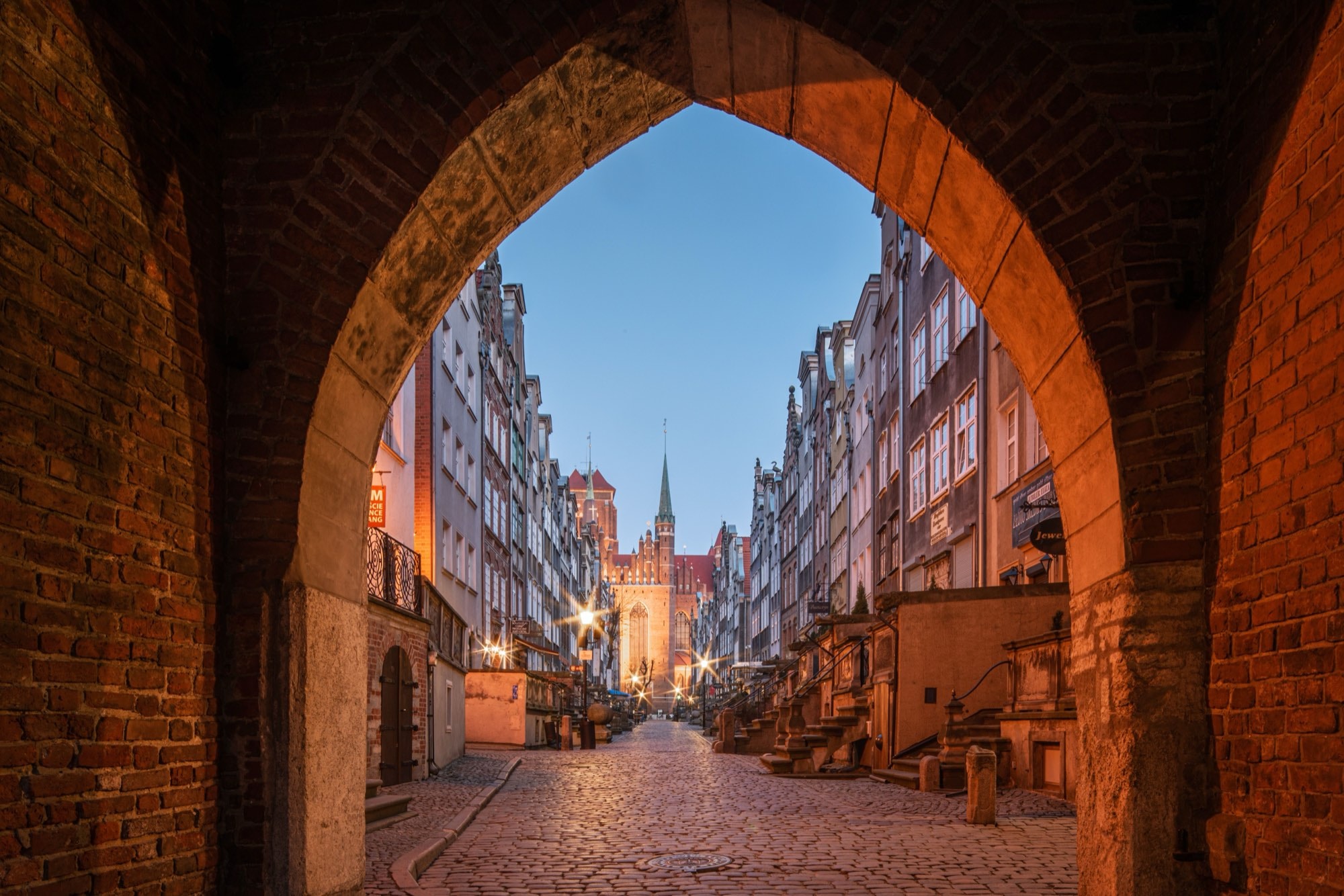 A brick Gothic gateway in Gdańsk's Old Town frames a cobbled lane at first light, street lamps warm against the tenement fronts and St Mary's closing the view