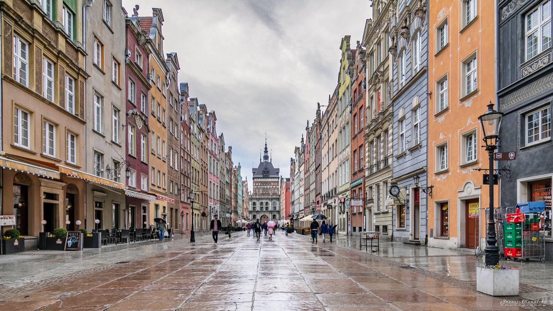 The Long Street — ulica Długa — in Gdańsk's Old Town after rain: colour-washed tenement fronts over wet flagstones, the Golden Gate closing the view, a few walkers in the distance